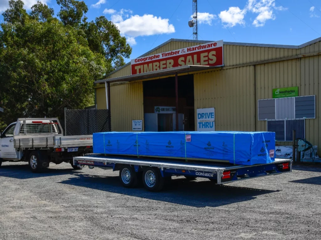 A truck with a trailer, loaded with timber wrapped in blue plastic, parked outside Geographe Timber & Hardware. The store's signage reads 'TIMBER SALES', and the clear sky and trees in the background complete the scene.