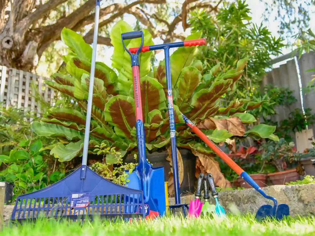 A selection of blue and red garden tools, including a plastic rake, digging spade, garden fork, and hoe, standing upright on a lawn next to potted plants.