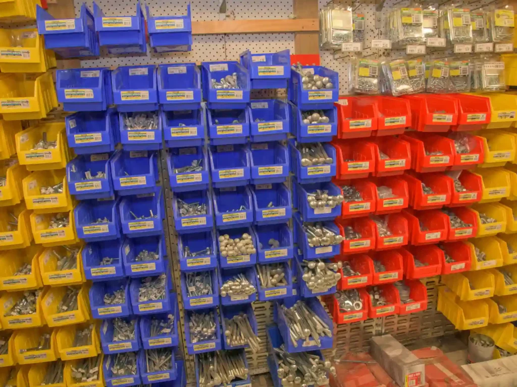 A pegboard wall display filled with yellow, blue, and red plastic storage bins holding assorted bulk bolts, screws, fasteners, and hardware fixings in store.