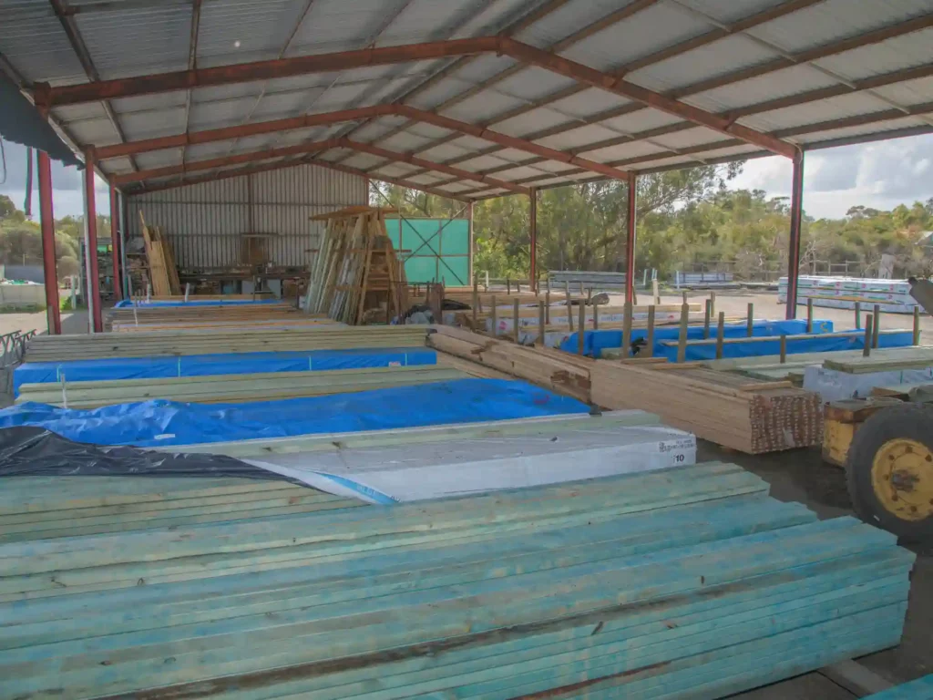 A wide interior view of a fully stocked commercial timber yard under a large open-sided steel warehouse shed, showing organized stacks of construction timber and framing lengths.