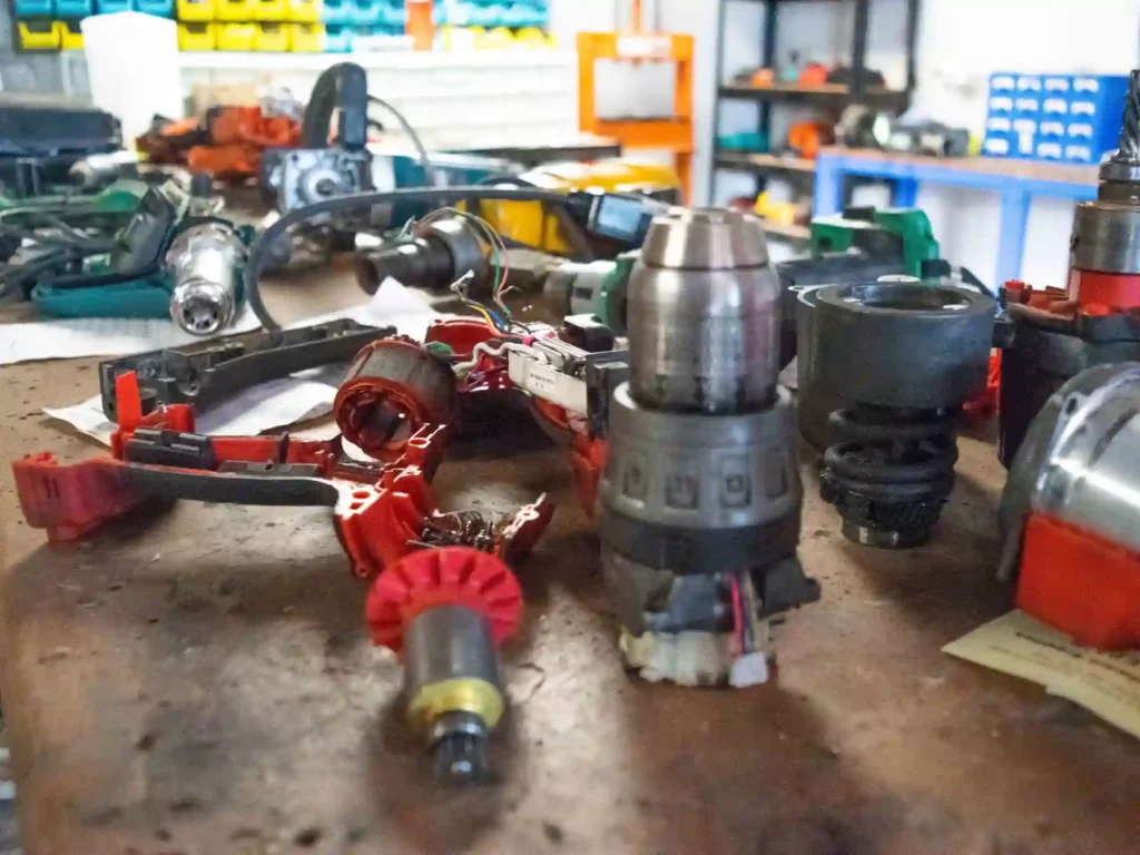 Disassembled components of cordless power tools, including a drill chuck, motor casing, and internal gears, laid out on a workshop bench for repairs.