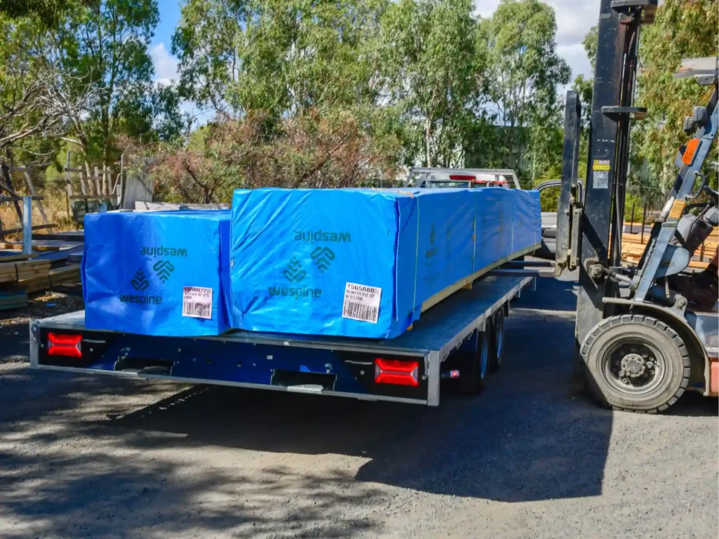 A heavy-duty warehouse forklift loading large timber packs wrapped in protective blue Wespine protective plastic covers onto a transport delivery trailer.