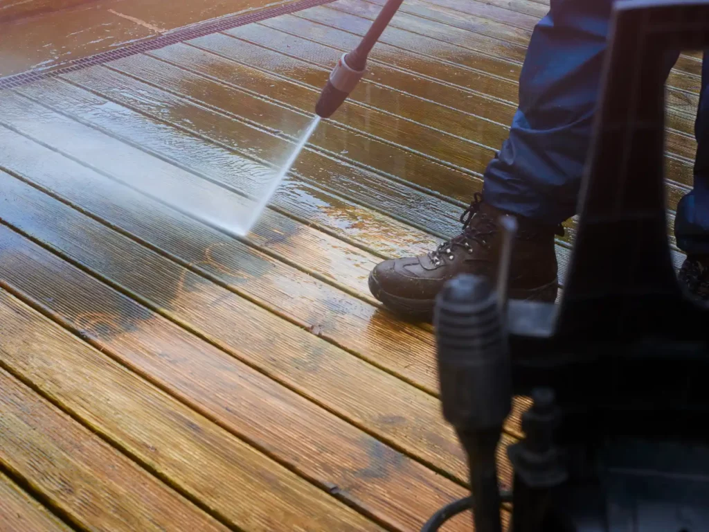 Close-up of a person using a high-pressure water cleaner to strip grime and clean a weathered outdoor timber deck panel.