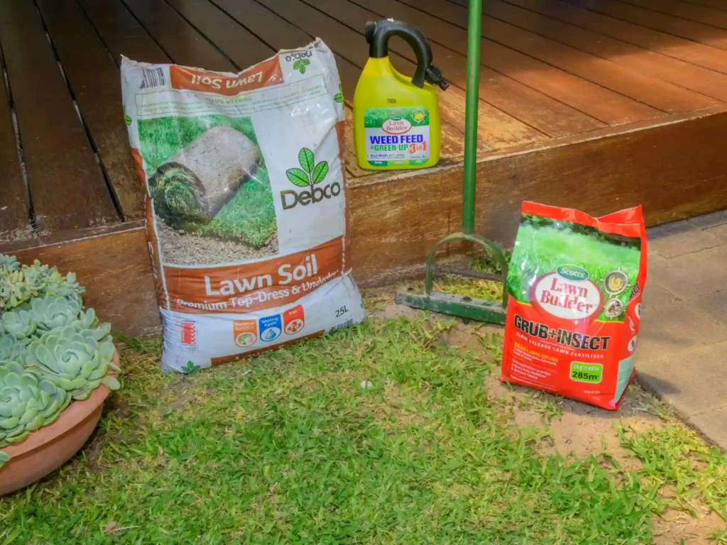A collection of spring lawn care products arrayed on a lawn next to a timber deck, including a bag of Debco Lawn Soil, Scotts Lawn Builder Weed Feed spray, and Scotts Grub Plus Insect control.