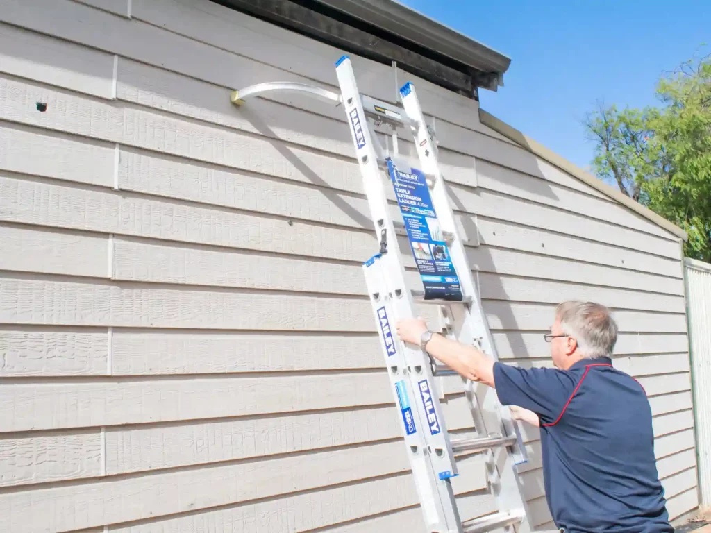 A man climbing a silver Bailey extension ladder propped up securely against the weatherboard siding of a house fascia to inspect the roof gutters.