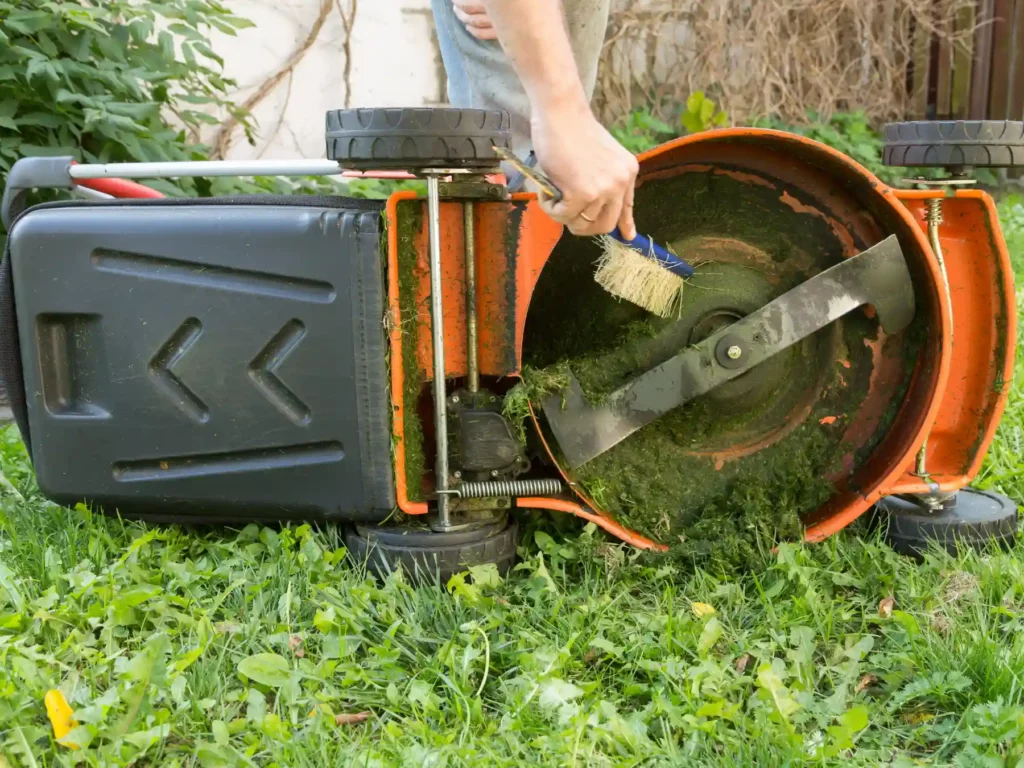 A person using a stiff-bristled brush to clear out packed green grass clippings from the underside blade chassis of an orange lawn mower sitting on a lawn.